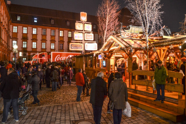 Menschen genießen einen winterlichen Abend im Bayreuther Winterdorf, mit festlich beleuchteten Holzhütten, geschmückten Bäumen und einem leuchtenden Coca-Cola-Weihnachtstruck im Hintergrund.
