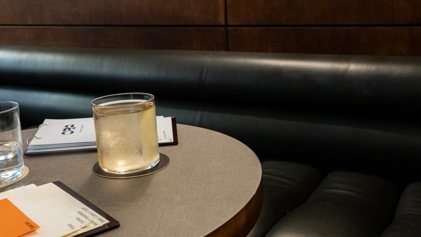 Detailaufnahme eines stilvollen Bartischs der Ora Bar in Zürich: ein Glas mit hellem Drink auf einem runden Tisch, daneben Wasserglas und Getränkekarten auf einer eleganten Lederbank – minimalistische, moderne Atmosphäre typisch für Zürcher Bars.