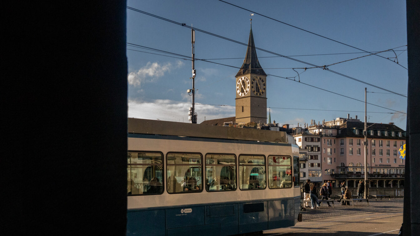 Straßenszene in Zürich mit einer vorbeifahrenden blauen Tram im Vordergrund und dem Turm der St. Peterskirche mit ihrer großen Uhr im Hintergrund, aufgenommen bei klarem Wetter am späten Nachmittag.