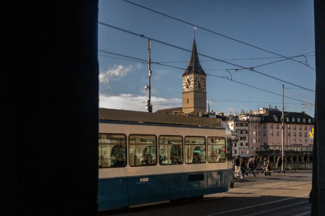 Straßenszene in Zürich mit einer vorbeifahrenden blauen Tram im Vordergrund und dem Turm der St. Peterskirche mit ihrer großen Uhr im Hintergrund, aufgenommen bei klarem Wetter am späten Nachmittag.