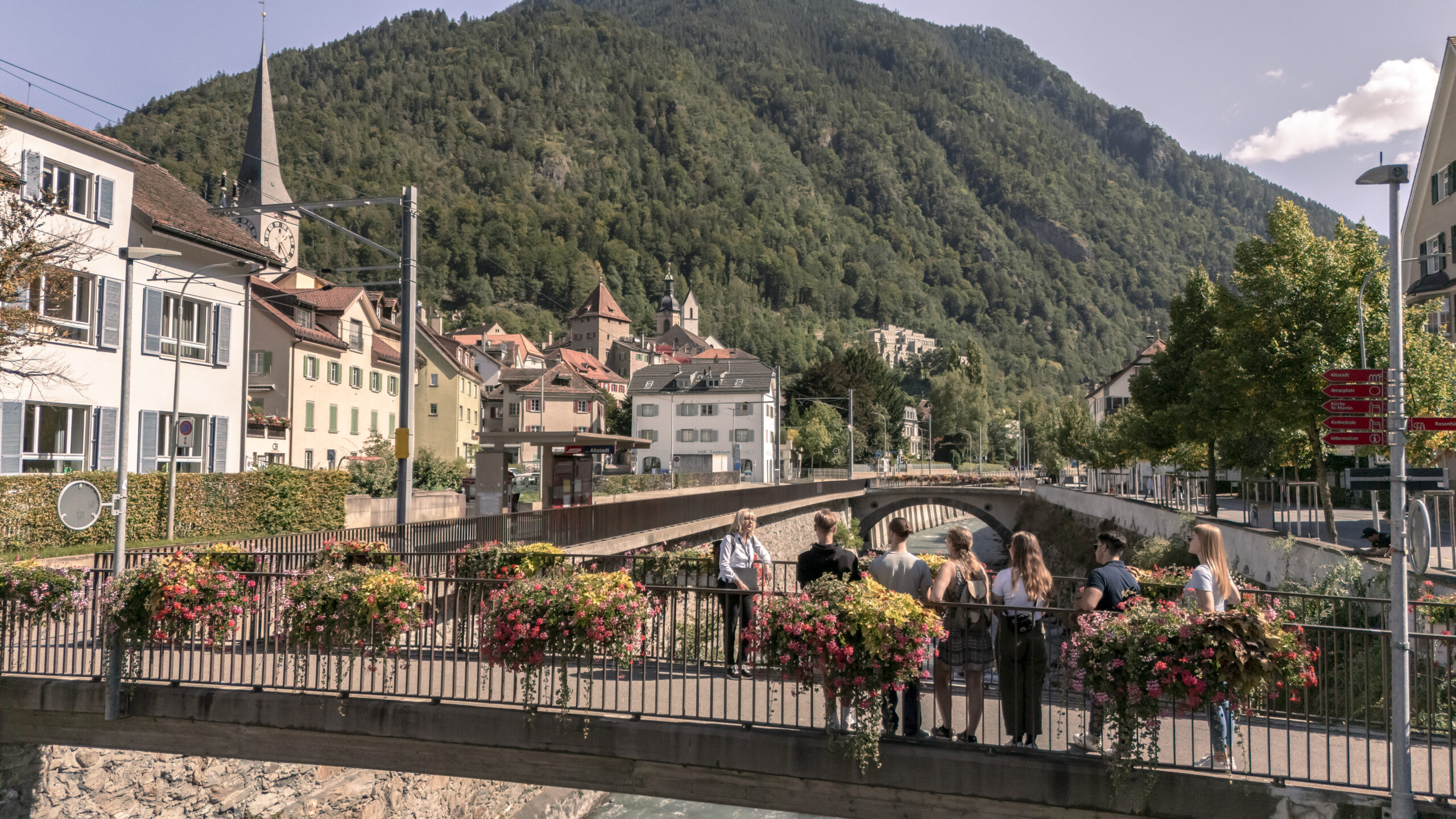 Eine Reisegruppe steht auf der Brücke der kleinen Schweizer Ortes Chur, im Hintergrund sieht man einen Berg.