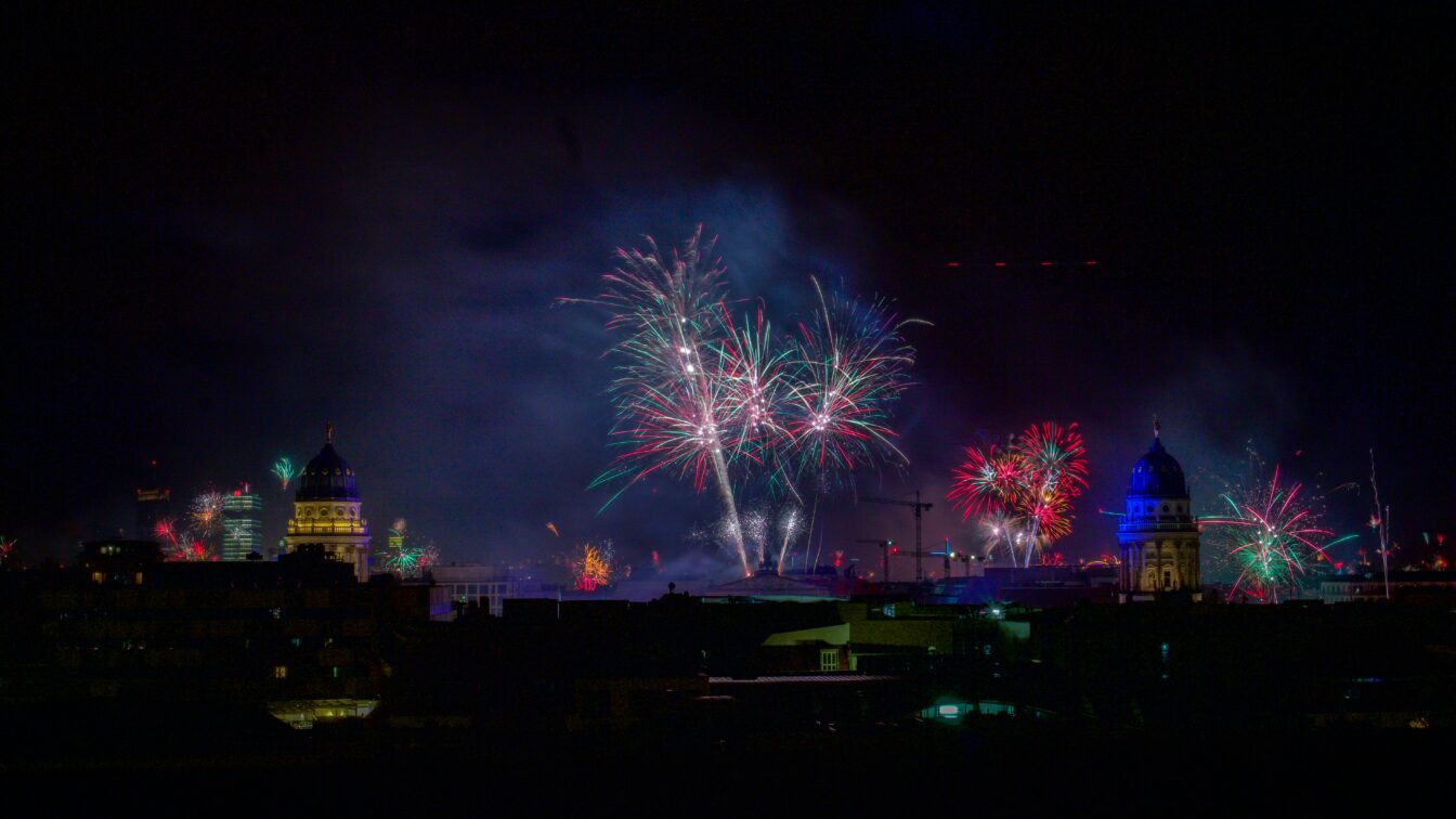 Nächtliche Skyline von Berlin an Silvester: Vor dunklem Himmel erhellen bunte Feuerwerksfontänen und Funkenexplosionen die Stadt. Links und rechts ragen zwei angestrahlte Kuppelbauten hervor, während im Hintergrund weitere Gebäude und Kräne schemenhaft sichtbar sind. Rauch und farbige Lichtspuren breiten sich über der Stadt aus.