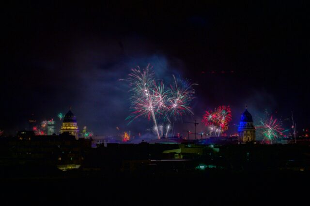 Nächtliche Skyline von Berlin an Silvester: Vor dunklem Himmel erhellen bunte Feuerwerksfontänen und Funkenexplosionen die Stadt. Links und rechts ragen zwei angestrahlte Kuppelbauten hervor, während im Hintergrund weitere Gebäude und Kräne schemenhaft sichtbar sind. Rauch und farbige Lichtspuren breiten sich über der Stadt aus.