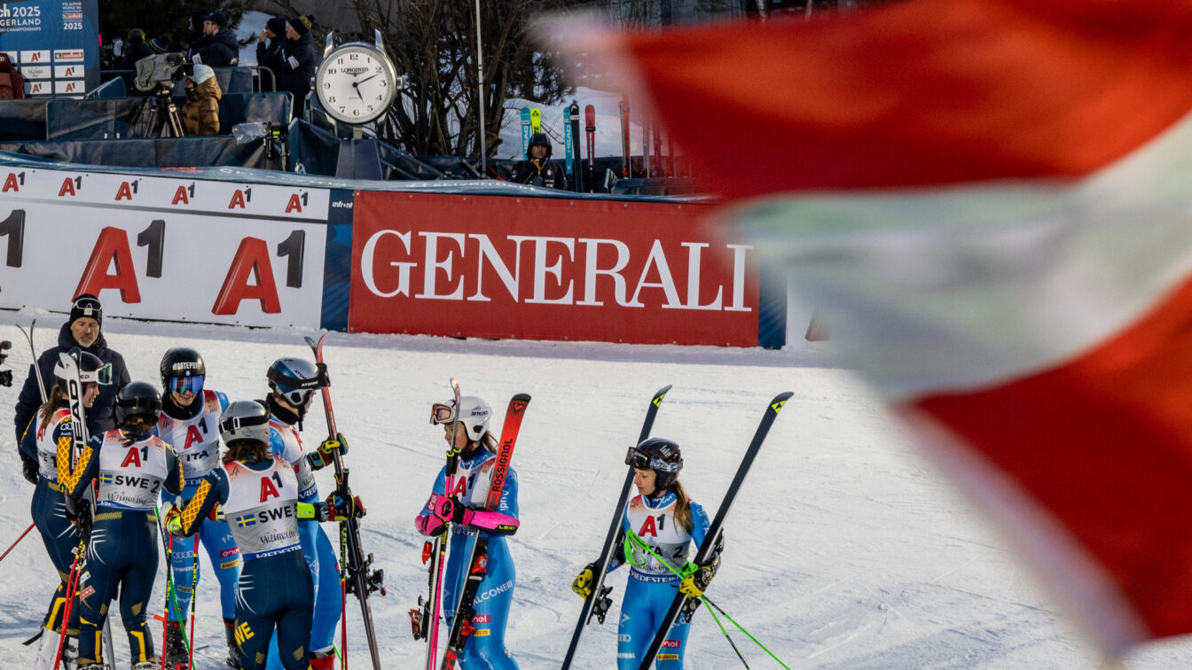 „Gruppe von Skirennläuferinnen in Rennanzügen und Helmen steht nach einem Wettkampf auf der Skipiste zusammen. Im Hintergrund sind Werbebanner, ein großer Standuhr und Zuschauer zu sehen; im Vordergrund weht unscharf eine rot-weiße Fahne ins Bild.