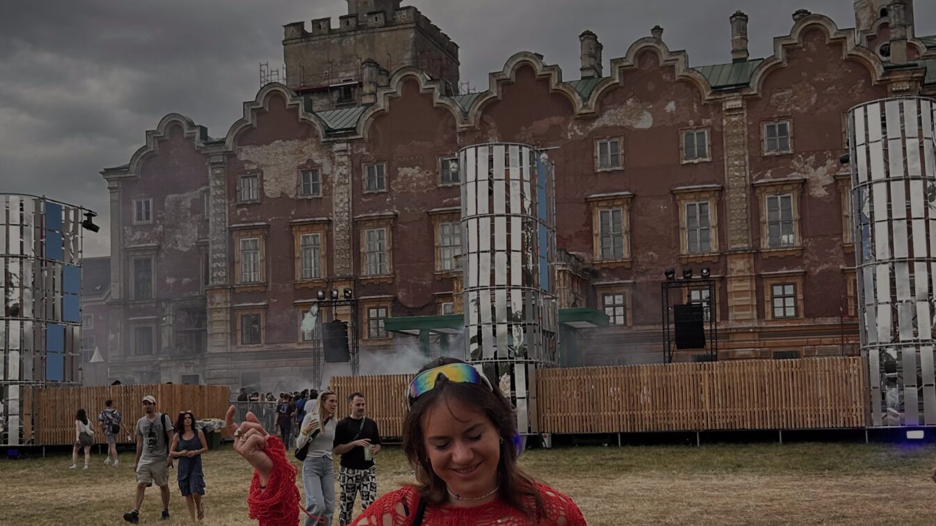 Person in rotem, grob gehäkeltem Oberteil tanzt lachend auf einem Open-Air-Techno-Festival vor einer verwitterten historischen Backsteinfassade; umgeben von Festivalbesucherinnen, Bühnenaufbauten und grauem, dramatischem Himmel.