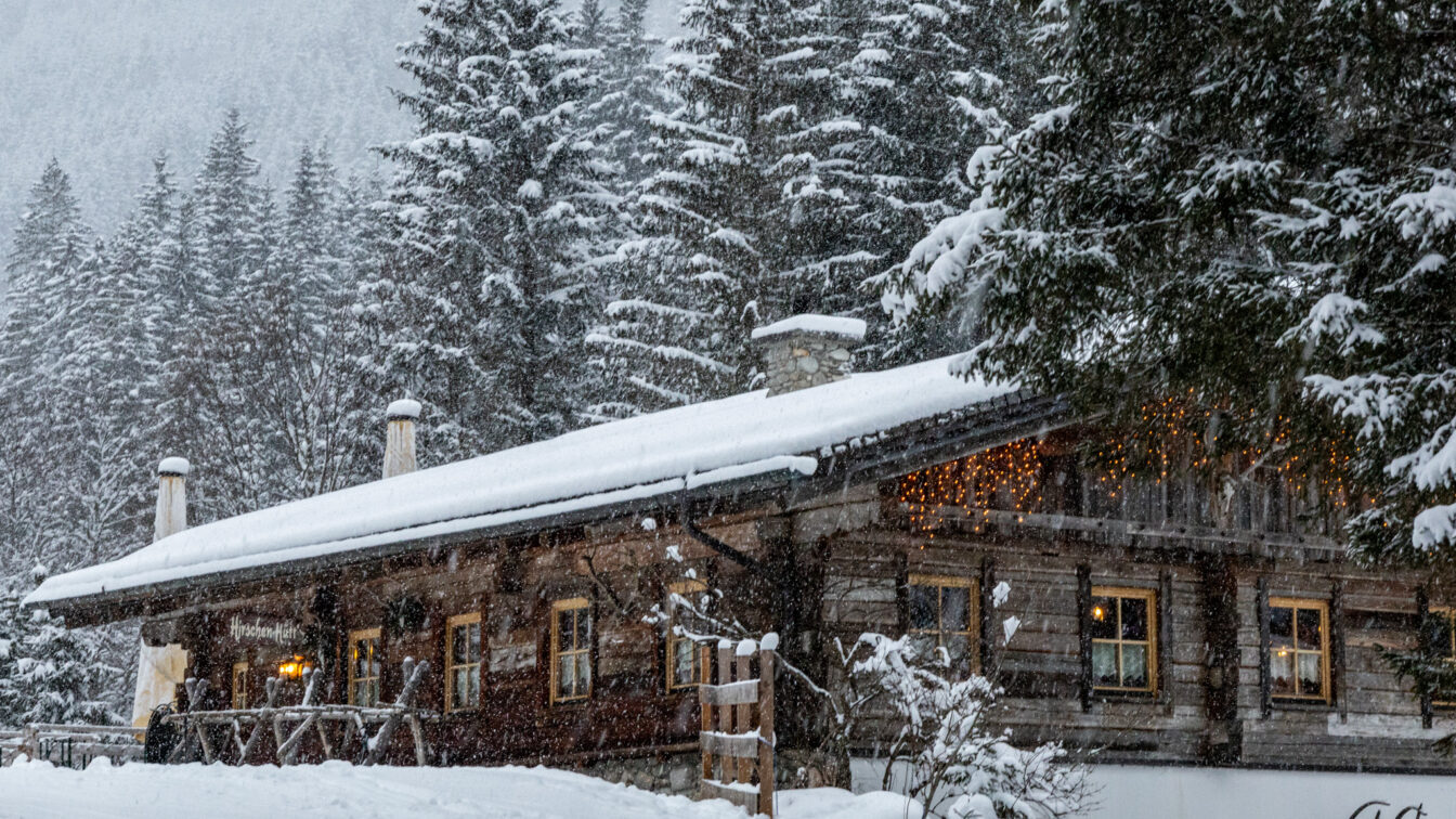 Ein verschneites Berghütten-Panorama in Österreich im Winter: Eine rustikale Holzhütte mit warm leuchtenden Fenstern und Lichterketten steht am Waldrand, umgeben von schneebedeckten Tannen und dicht fallendem Schnee.
