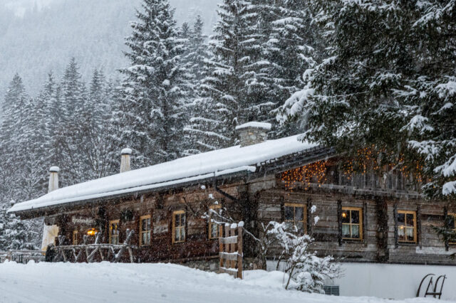 Ein verschneites Berghütten-Panorama in Österreich im Winter: Eine rustikale Holzhütte mit warm leuchtenden Fenstern und Lichterketten steht am Waldrand, umgeben von schneebedeckten Tannen und dicht fallendem Schnee.