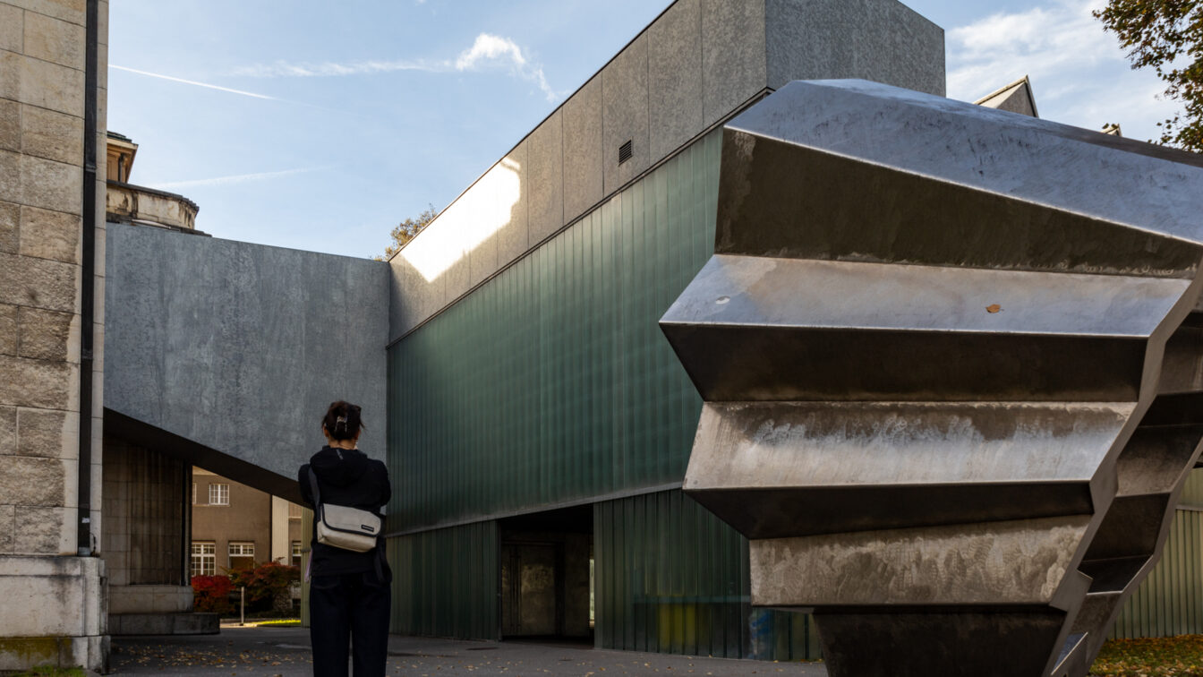 Eine Person steht vor einem modernen Museumsgebäude mit klaren geometrischen Formen, grünlich getönten Glaswänden und einer markanten Metallskulptur im Vordergrund. Das Wechselspiel aus Stein, Glas und Metall verleiht dem Ort eine architektonisch spannende Atmosphäre. Das Bild zeigt Winterthur, der sich gut als Ausflugsziel ab Zürich eignet.