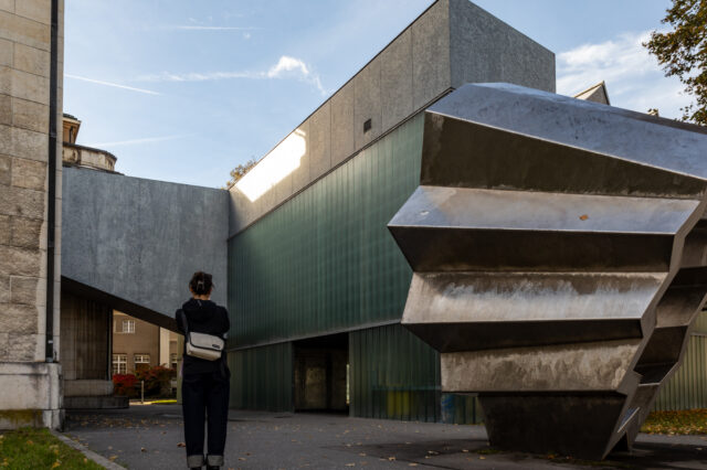 Eine Person steht vor einem modernen Museumsgebäude mit klaren geometrischen Formen, grünlich getönten Glaswänden und einer markanten Metallskulptur im Vordergrund. Das Wechselspiel aus Stein, Glas und Metall verleiht dem Ort eine architektonisch spannende Atmosphäre. Das Bild zeigt Winterthur, der sich gut als Ausflugsziel ab Zürich eignet.