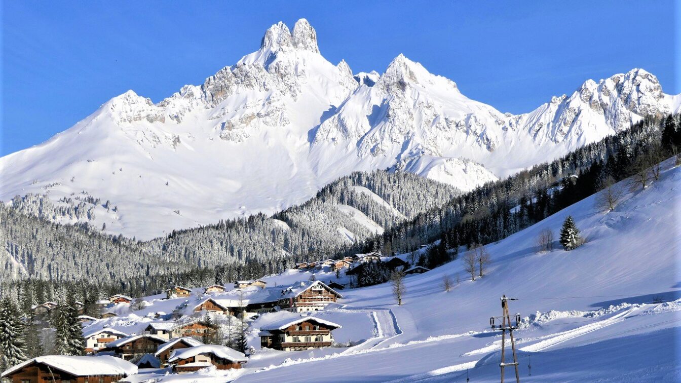 Auf dem Bild sieht man das verschneite Bergdorf Filzmoos mit zahlreichen Holzhäusern, eingebettet in eine Winterlandschaft, überragt vom markanten, schneebedeckten Gipfel der Bischofsmütze.