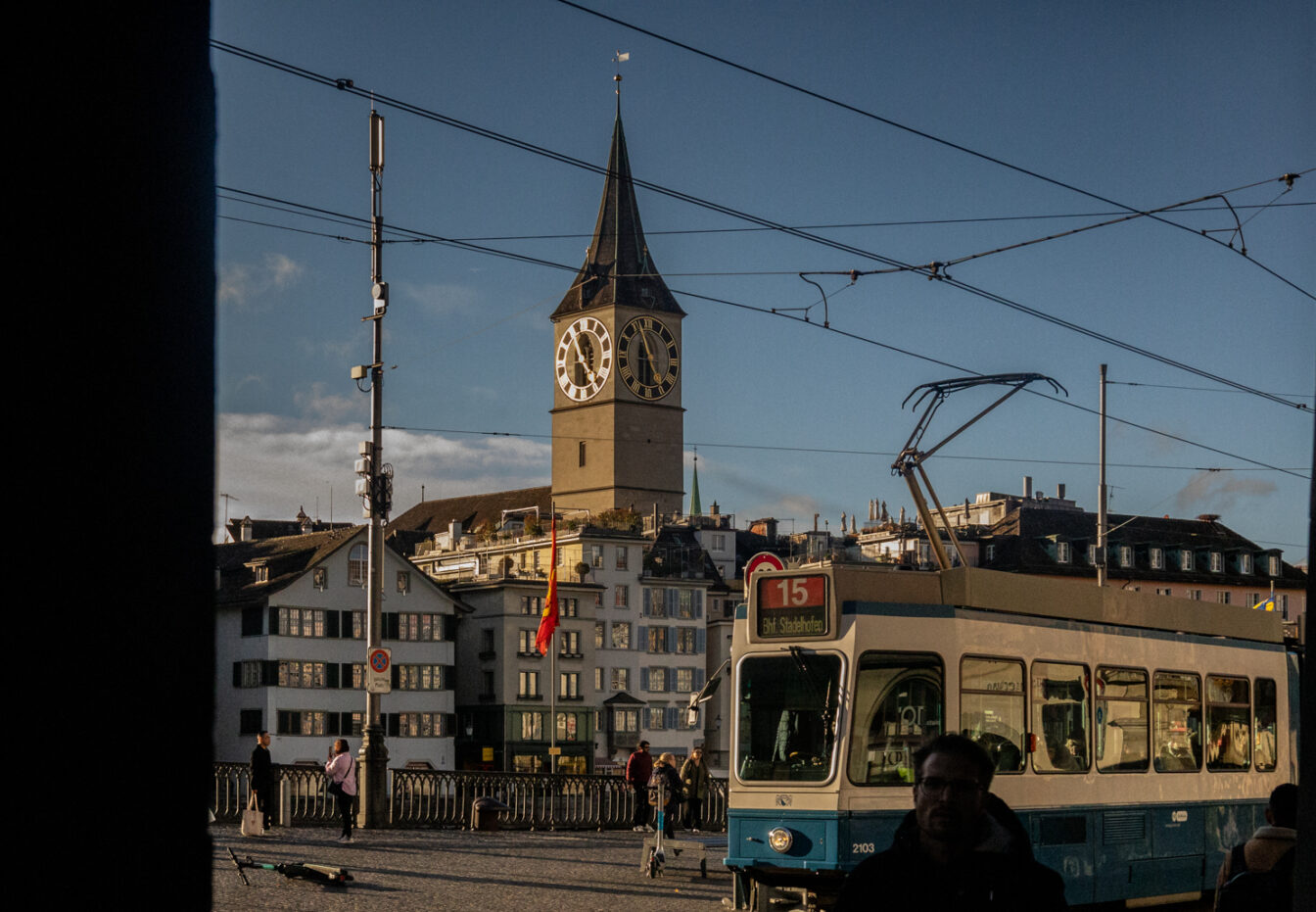 Straßenszene in Zürich bei tiefstehender Sonne: Eine blau-weiße Tram der Linie 15 fährt rechts ins Bild, während Menschen auf dem Platz unterwegs sind. Im Hintergrund steht der markante Turm der St.-Peter-Kirche mit seiner großen Uhr. Dahinter reihen sich traditionelle Altstadthäuser. Stromleitungen für die Tram durchziehen den Himmel.