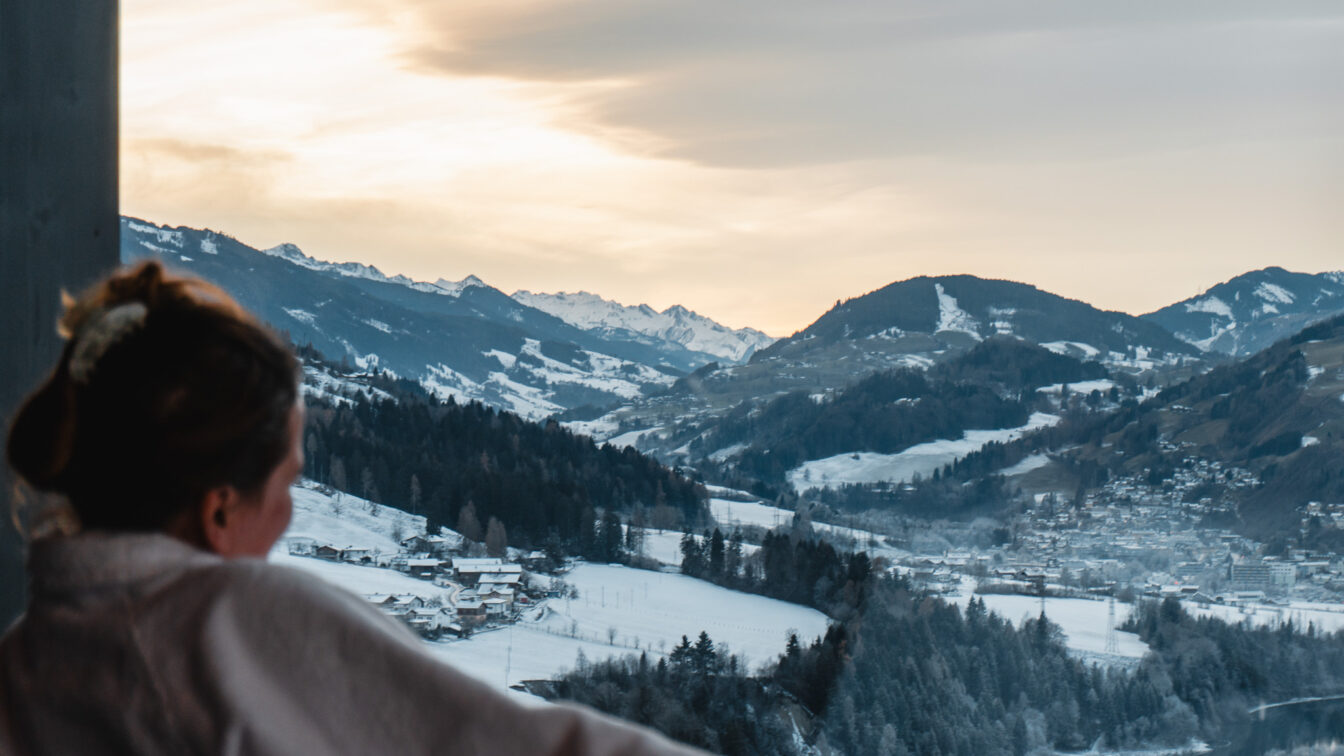 Ausblick von einem Hotelzimmer auf verschneite Alpenlandschaft mit Skipisten, Bergdorf und winterlichen Wäldern bei Sonnenaufgang