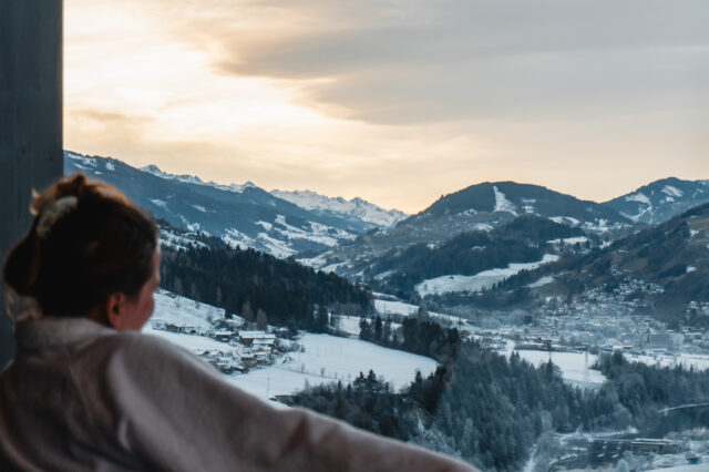 Ausblick von einem Hotelzimmer auf verschneite Alpenlandschaft mit Skipisten, Bergdorf und winterlichen Wäldern bei Sonnenaufgang