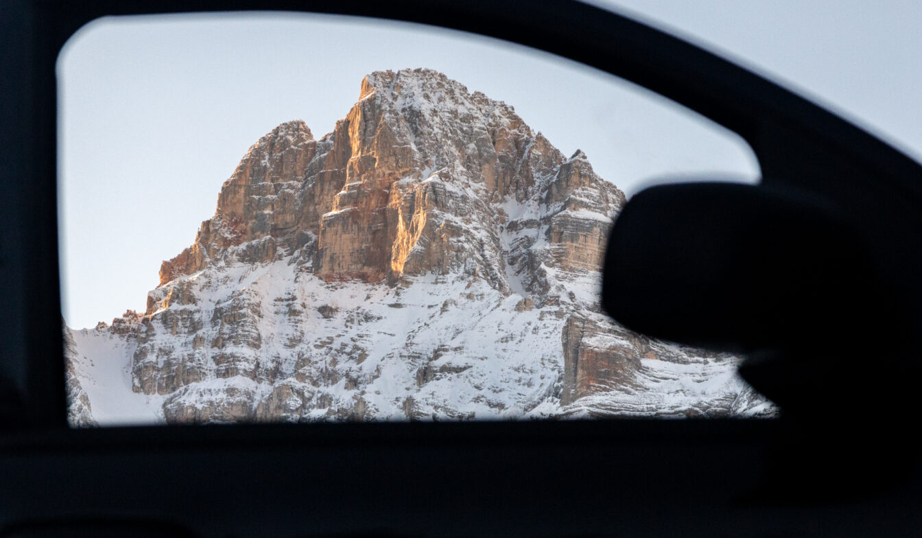 Schneebedeckter Berggipfel in warmem Morgen- oder Abendlicht, durch den Rahmen eines geöffneten Autofensters fotografiert; der dunkle Innenraum des Autos bildet einen Kontrast zum hellen Felsmassiv