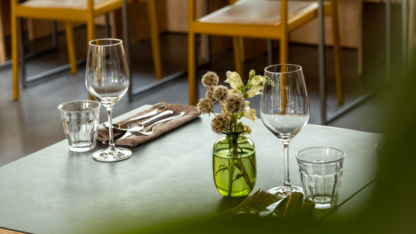 „Elegant gedeckter Restauranttisch mit zwei Weingläsern, Wassergläsern, Besteck auf einer braunen Serviette und einer kleinen Vase mit weißen Blumen; im Hintergrund gelbe Stühle in einem modernen Innenraum.“