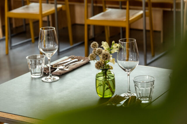 „Elegant gedeckter Restauranttisch mit zwei Weingläsern, Wassergläsern, Besteck auf einer braunen Serviette und einer kleinen Vase mit weißen Blumen; im Hintergrund gelbe Stühle in einem modernen Innenraum.“