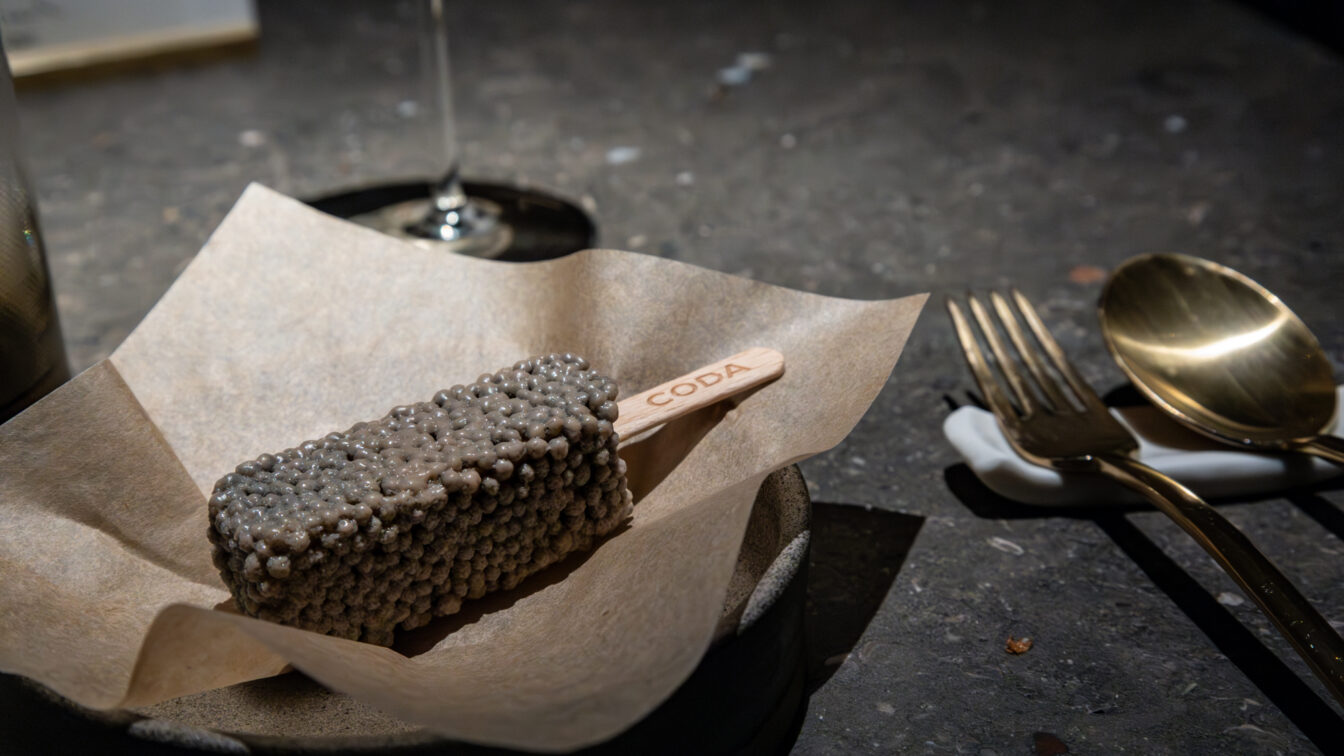 Gourmet-Dessert am Stiel im Fine-Dining-Restaurant in Berlin: ein rechteckiges Eis mit perlartiger Textur auf braunem Papier serviert, daneben elegantes goldenes Besteck