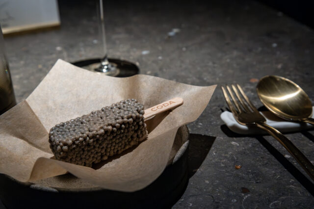 Gourmet-Dessert am Stiel im Fine-Dining-Restaurant in Berlin: ein rechteckiges Eis mit perlartiger Textur auf braunem Papier serviert, daneben elegantes goldenes Besteck