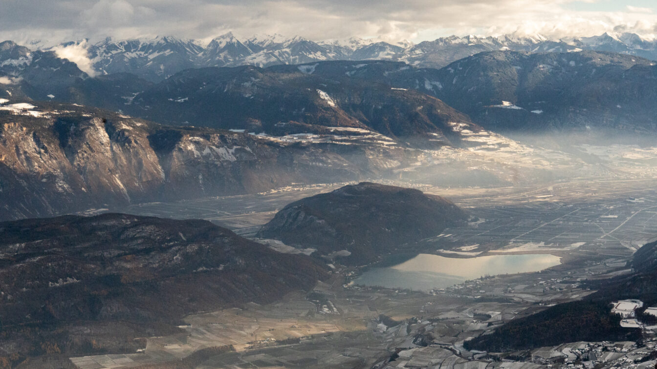 Weitwinkelaufnahme des Etschtals mit dem Kalterer See und umliegenden Hügeln im Vordergrund; dahinter breite Bergketten mit Schnee und teils diffusem Licht.