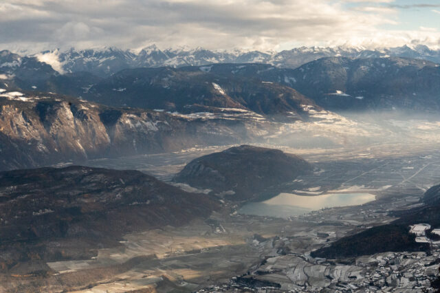 Weitwinkelaufnahme des Etschtals mit dem Kalterer See und umliegenden Hügeln im Vordergrund; dahinter breite Bergketten mit Schnee und teils diffusem Licht.