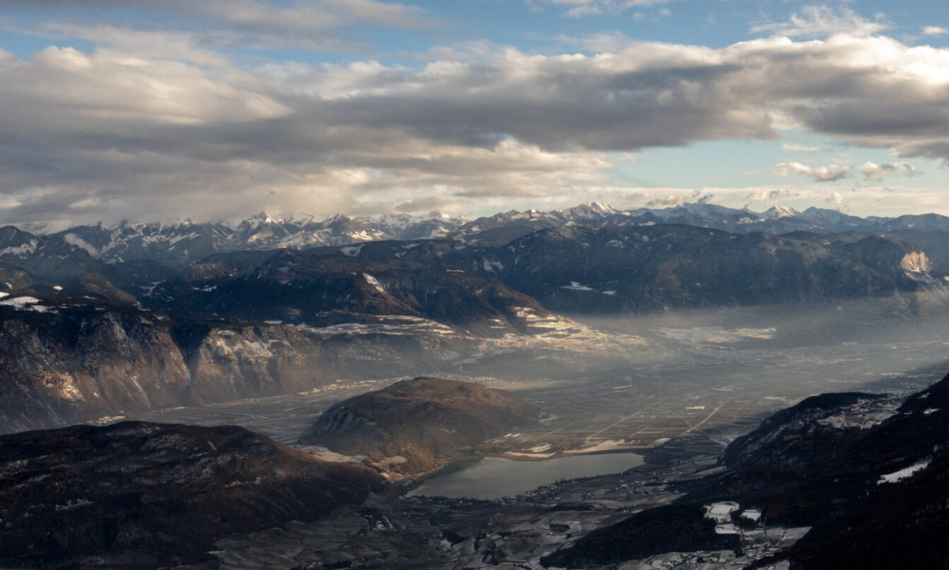 Panorama des Etschtals mit sichtbarem Kalterer See, Hügeln und Bergketten im Hintergrund, unter einem bewölkten, teilweise aufgehellten Himmel.