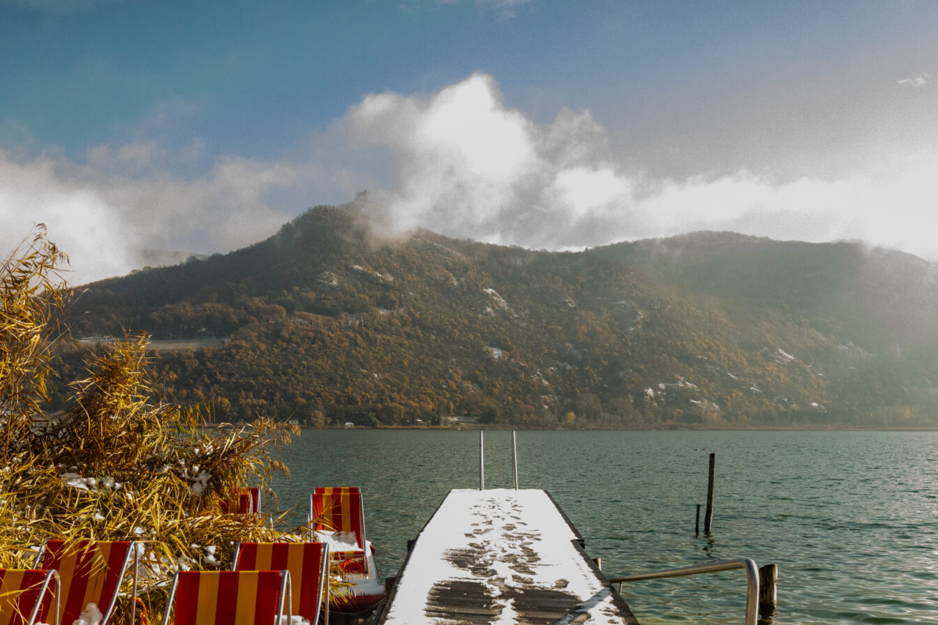 Schmaler verschneiter Steg mit Spuren im Schnee, rechts und links bunte Liegestühle, dahinter der Kalterer See und bewaldete Hügel mit etwas Schnee.
