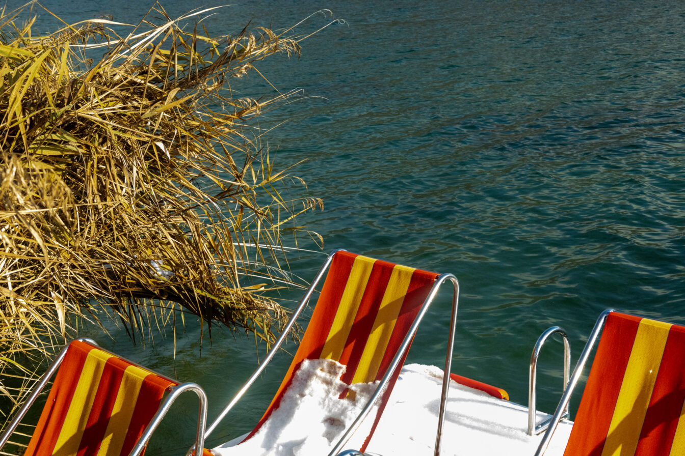 Nahaufnahme eines orange-gelb gestreiften Liegestuhls mit Schnee darauf, daneben Schilf am Ufer und das türkisblaue Wasser des Kalterer Sees.