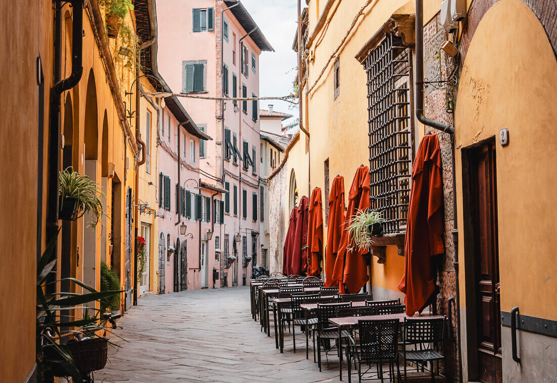 Schmale, gepflasterte Gasse in einer italienischen Altstadt mit warm gelb- und rosafarbenen Hausfassaden; rechts stehen leere Café-Tische mit geschlossenen roten Sonnenschirmen, links lehnt ein Fahrrad an der Wand. Die Gasse führt leicht gebogen auf weitere historische Gebäude zu, überragt von einem Bogen im Vordergrund.