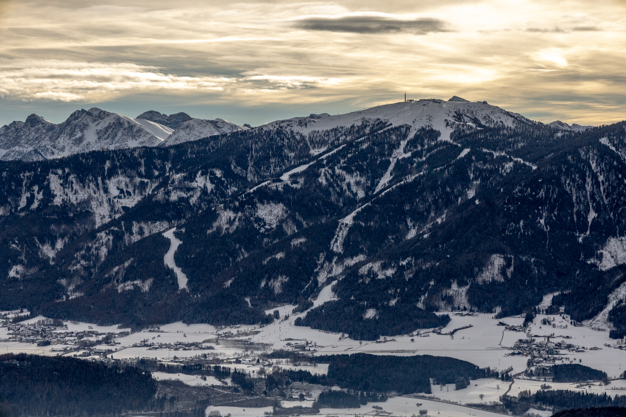 Winterliche Berglandschaft im Pustertal mit schneebedeckten Hängen, dunklen Waldflächen und markanten Gipfeln im Hintergrund. Im Tal liegen verstreute Dörfer und Felder unter einer dünnen Schneedecke. Der Himmel ist von sanftem Abend- oder Morgengold beleuchtet und teils von Wolken überzogen