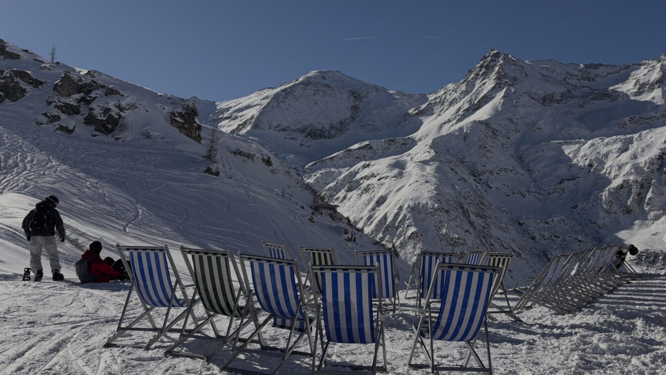 Eine winterliche Berglandschaft mit schneebedeckten Gipfeln unter blauem Himmel. Im Vordergrund stehen mehrere gestreifte Liegestühle im Schnee, daneben zwei Wintersportler*innen mit Ausrüstung. Die Szene wirkt sonnig, ruhig und einladend.