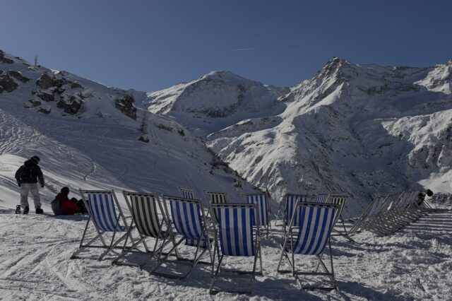 Eine winterliche Berglandschaft mit schneebedeckten Gipfeln unter blauem Himmel. Im Vordergrund stehen mehrere gestreifte Liegestühle im Schnee, daneben zwei Wintersportler*innen mit Ausrüstung. Die Szene wirkt sonnig, ruhig und einladend.