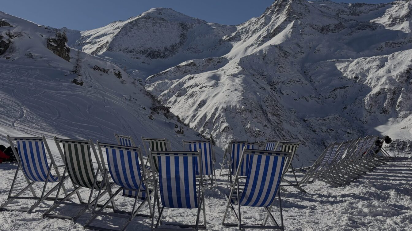 Eine winterliche Berglandschaft mit schneebedeckten Gipfeln unter blauem Himmel. Im Vordergrund stehen mehrere gestreifte Liegestühle im Schnee, daneben zwei Wintersportler*innen mit Ausrüstung. Die Szene wirkt sonnig, ruhig und einladend.