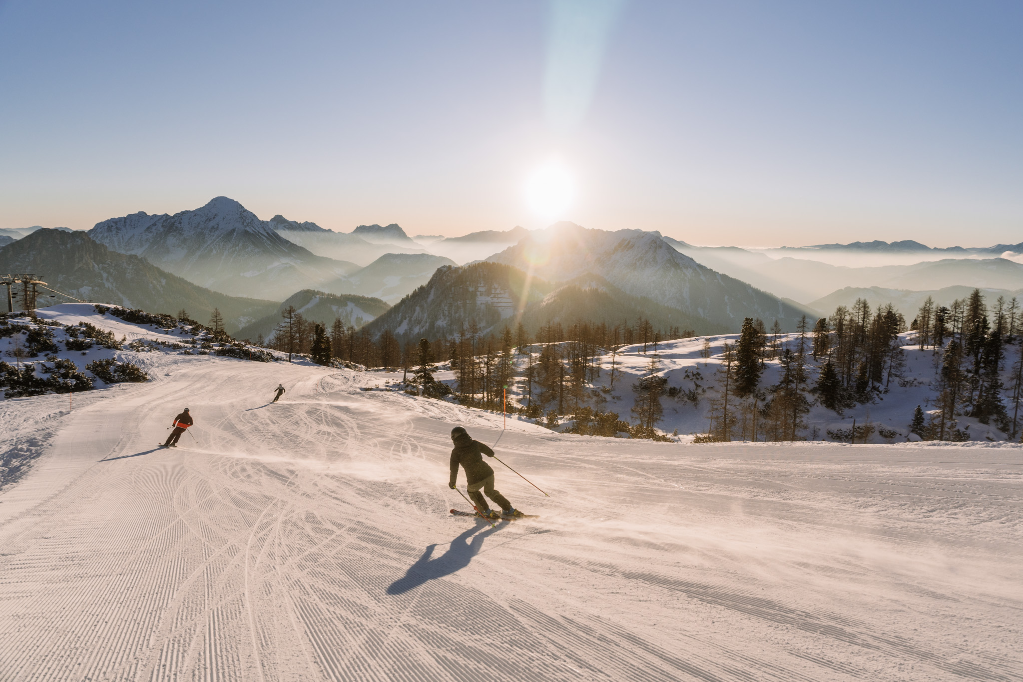 Eine Person fährt eine verschneite Piste herunter, in der Ferne ist eine malerische und wolkenbedeckte Gebirgskette und die untergehende Sonne zu sehen.