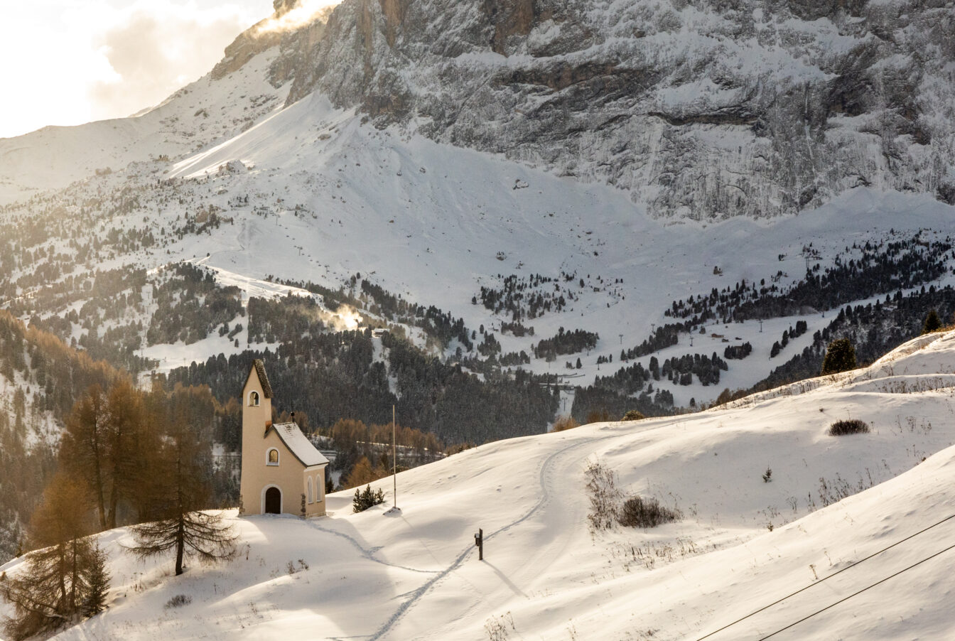 Winterlandschaft in Südtirol mit einer kleinen Kapelle auf verschneiten Hügeln und den Dolomiten im Hintergrund.