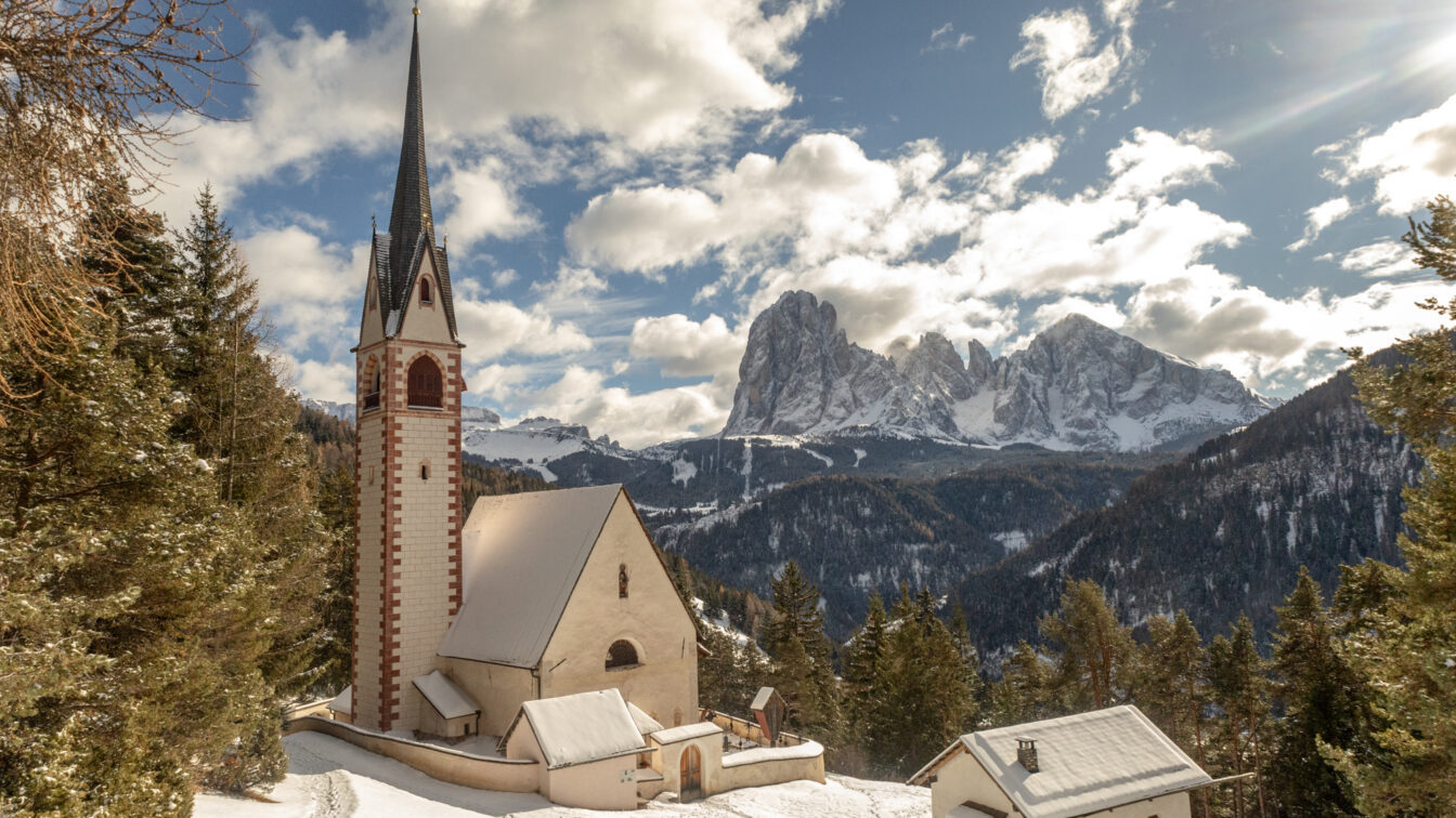 Winterlandschaft in Südtirol mit der verschneiten St.-Jakobs-Kirche auf einer Anhöhe des Grödnertals. Vor dem Panorama ragen die schneebedeckten Gipfel der Geislergruppe in den Himmel. Rund um die Kirche liegen Waldhänge und einzelne Höfe, während Sonnenstrahlen durch die Wolken brechen und die Szene warm beleuchten.