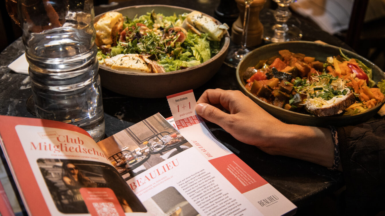 Zwei angerichtete Gerichte auf einem Restauranttisch, daneben ein aufgeschlagenes Genussheft mit Gutschein und eine Hand, die einen 1+1-Voucher hält.
