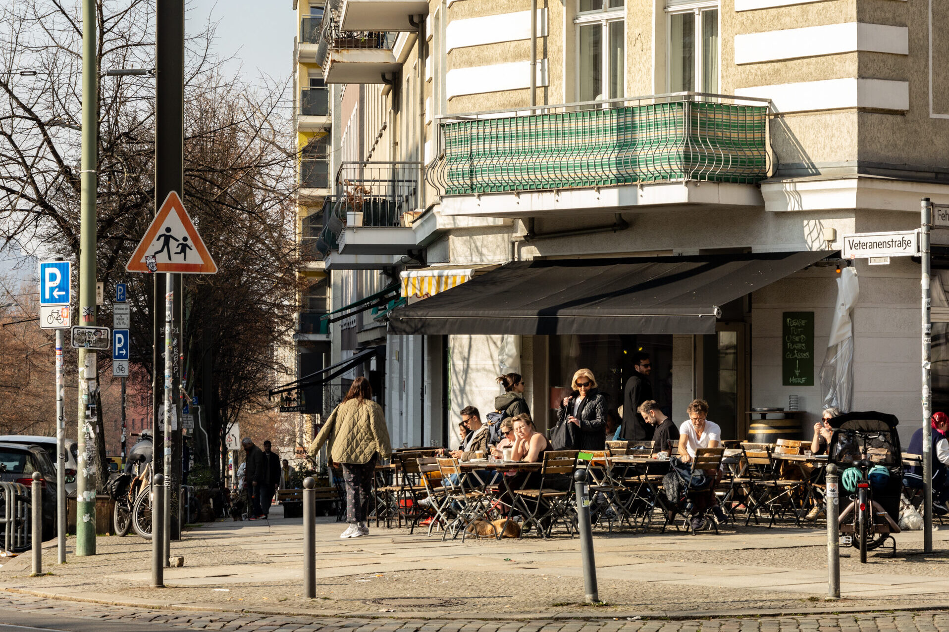 Ein typischer Berliner Straßenzug mit einer einladenden Café- und Bar-Szene. Die Szene spielt sich an der Veteranenstraße ab, wo Menschen das sonnige Wetter genießen und sich an den Tischen eines Straßencafés niederlassen. Schwarze Markisen spenden Schatten, während die Gäste in entspannter Atmosphäre sitzen, plaudern und ihr Essen oder Getränke genießen.