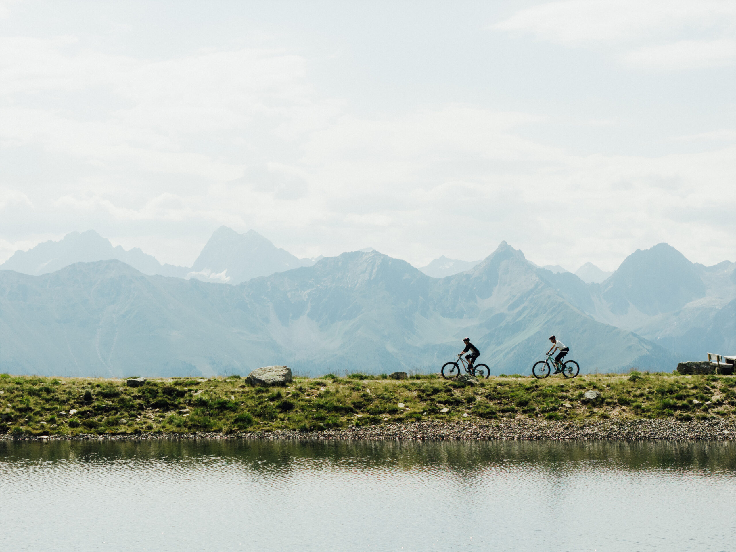 Zwei Radfahrer fahren an einem See entlang, dahinter ist ein vernebeltes Bergpanorama zu sehen.