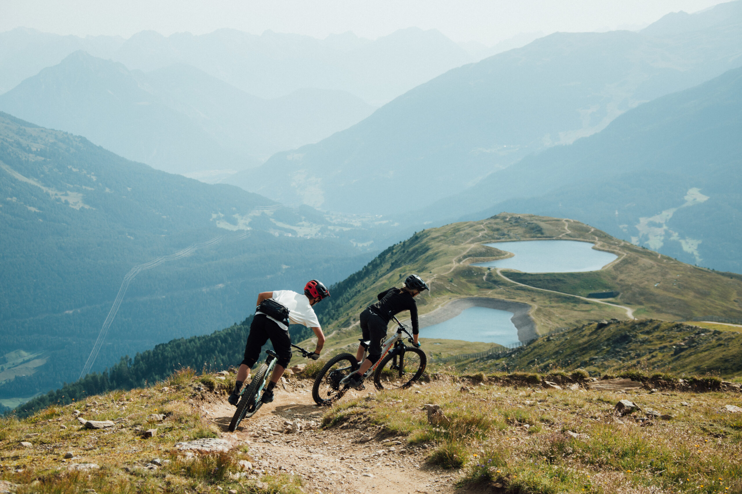 Zwei Radfahrer fahren am Frommestrail in Serfaus-Fiss-Ladis einen Trail entlang auf zwei kleine Seen zu, im Hintergrund ist ein Bergpanorama zu sehen.