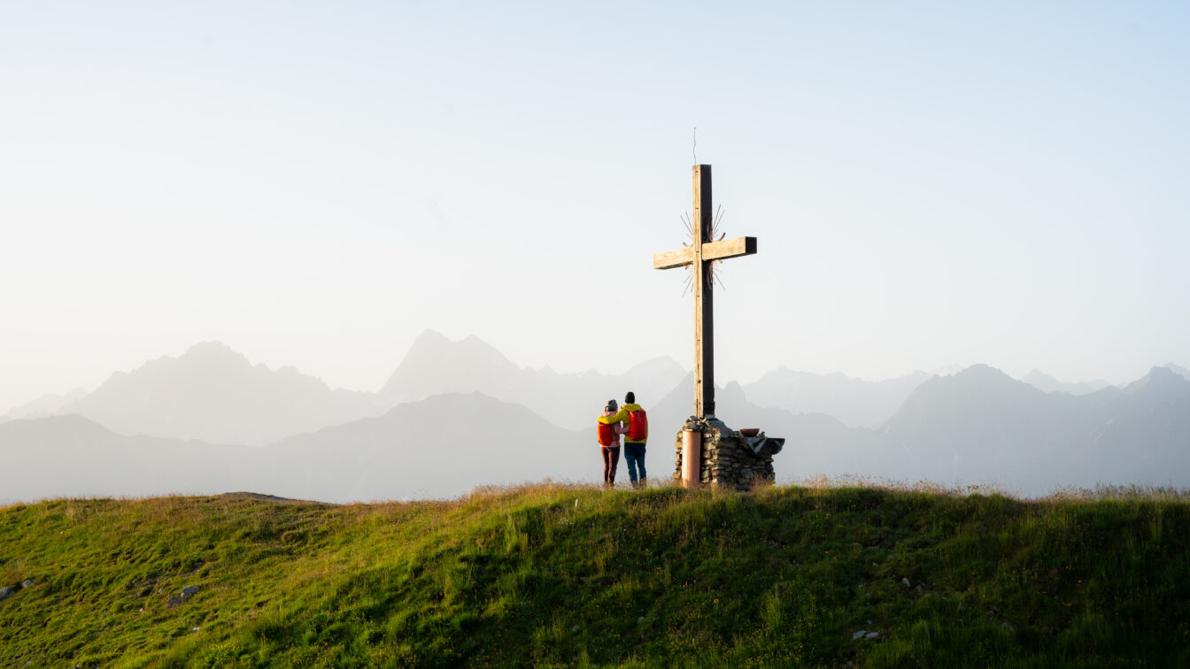Zwei Personen stehen auf einem Berg beim Gipfelkreuz und bewundern die Berglandschaft.