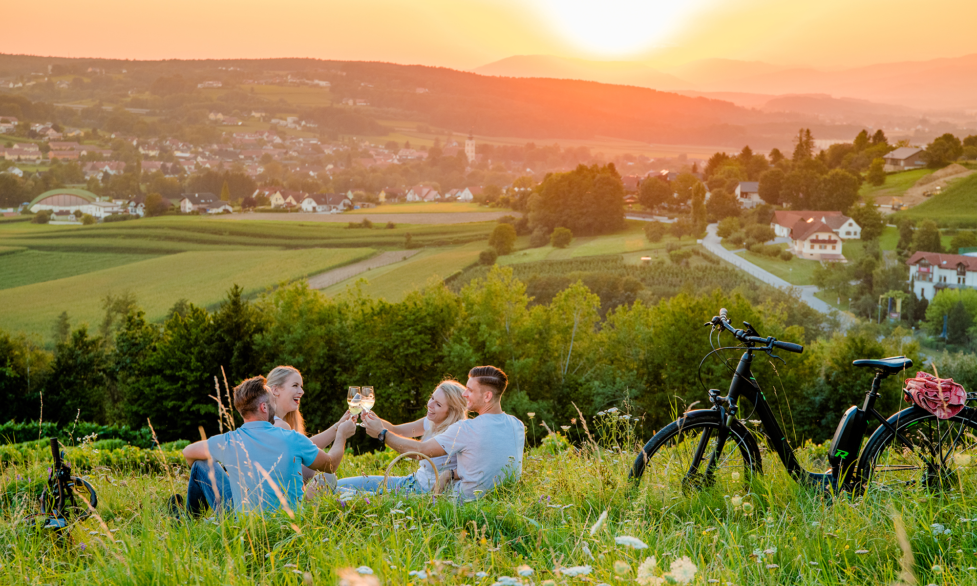 Hügelige Landschaft mit Weingarten bei Sonnenuntergang. Im Vordergrund stoßt eine Gruppe von Menschen mit Wiengläsern an.
