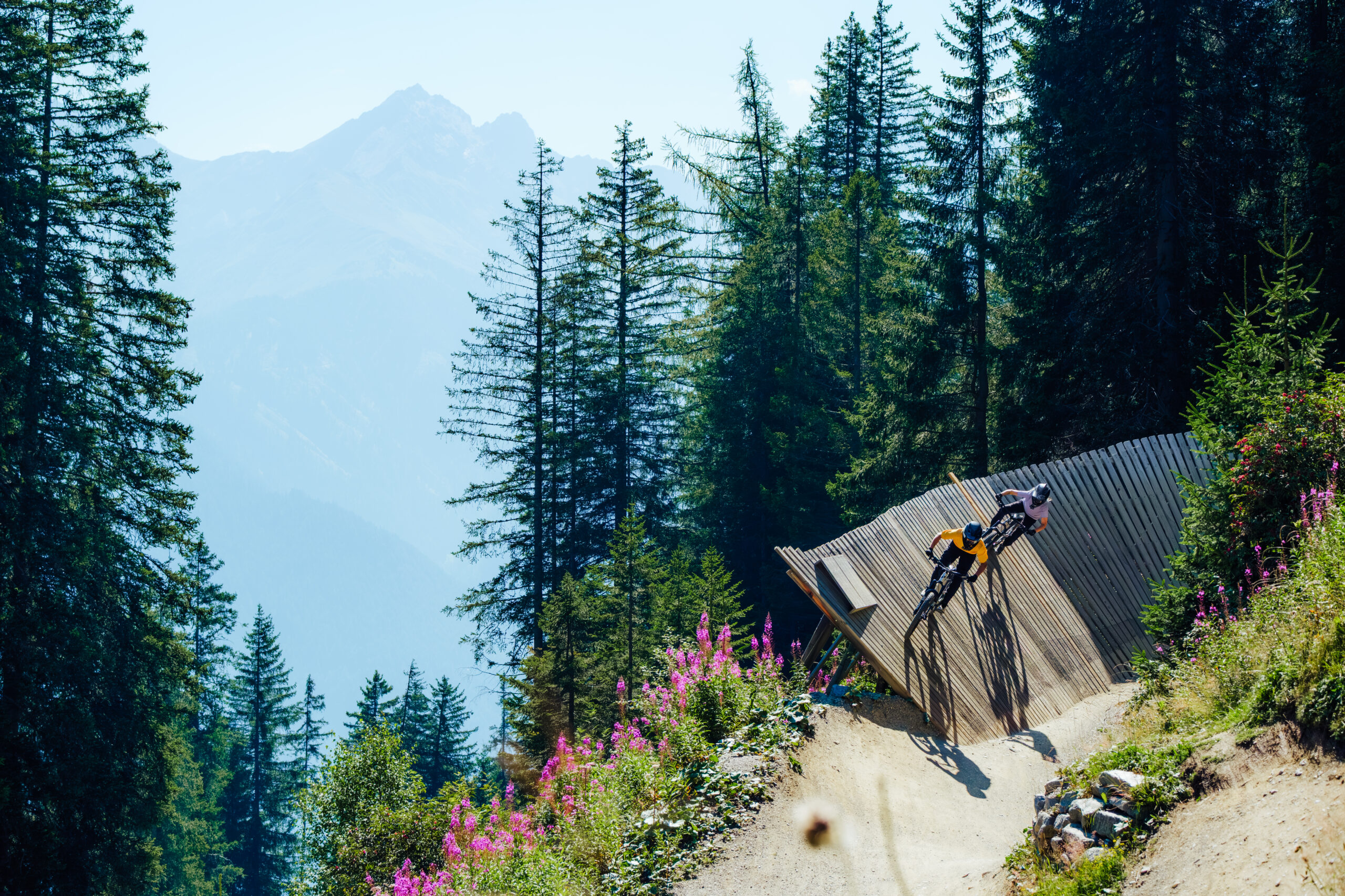 Zwei Radfahrer fahren im Bikepark Serfaus-Fiss-Ladis auf einem Steg um die Kurve, im Hintergrund ist ein Bergpanorama zu sehen.