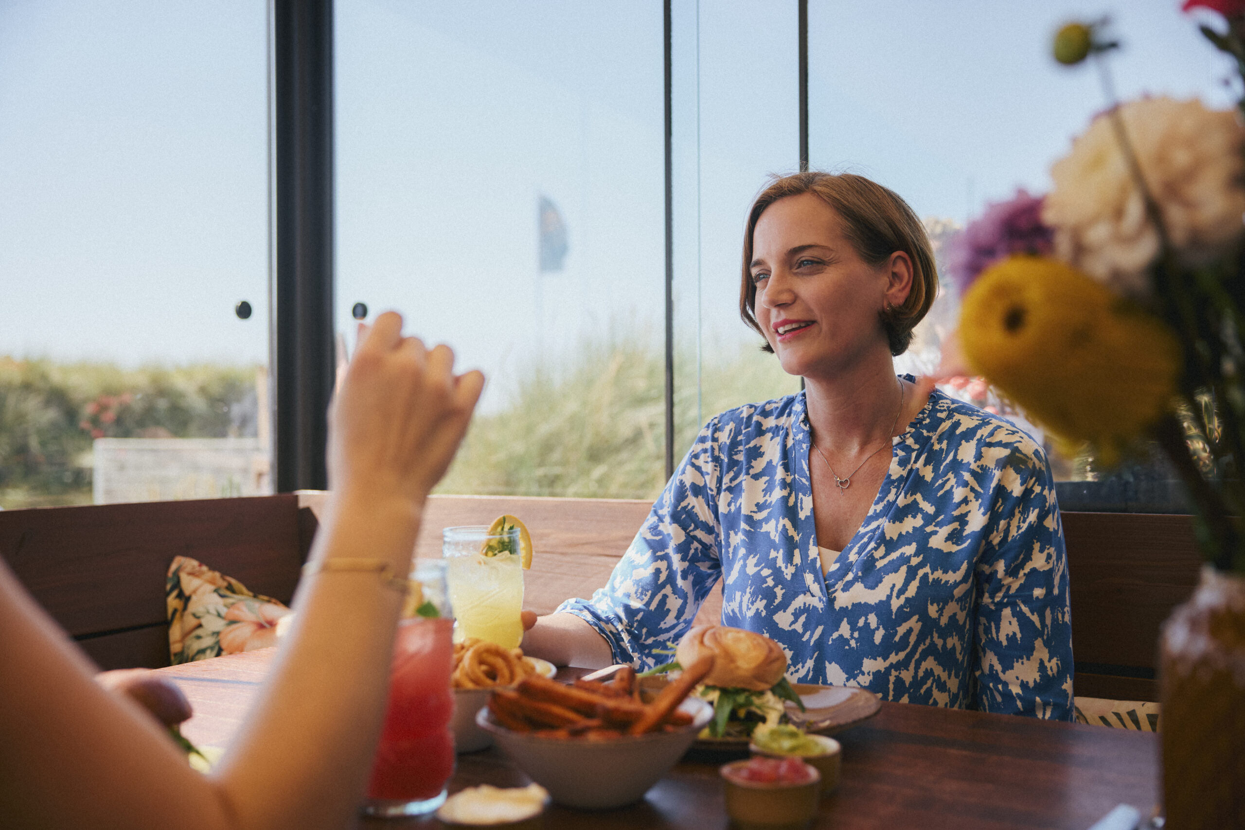 Zwei Frauen sitzen im Twisters Beach Diner in Wenningstedt auf Sylt an einem Holztisch mit einer Auswahl an Speisen und Cocktails. Die Frau im Fokus trägt ein blau-weißes Kleid und führt eine lebhafte Unterhaltung, während draußen durch große Fenster die Natur zu sehen ist.