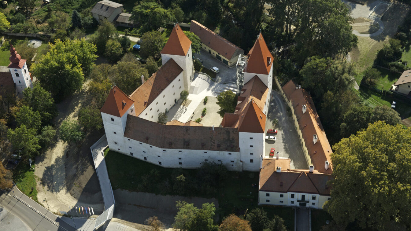 Luftaufnahme von Schloss Orth: Weiße Schlossanlage mit vier markanten roten Spitztürmen, umgeben von Bäumen.