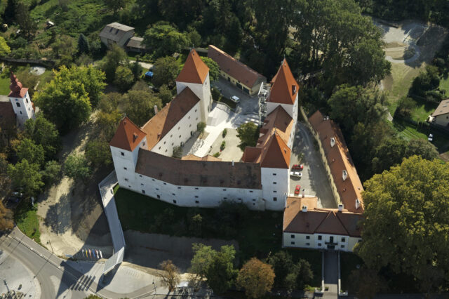 Luftaufnahme von Schloss Orth: Weiße Schlossanlage mit vier markanten roten Spitztürmen, umgeben von Bäumen.