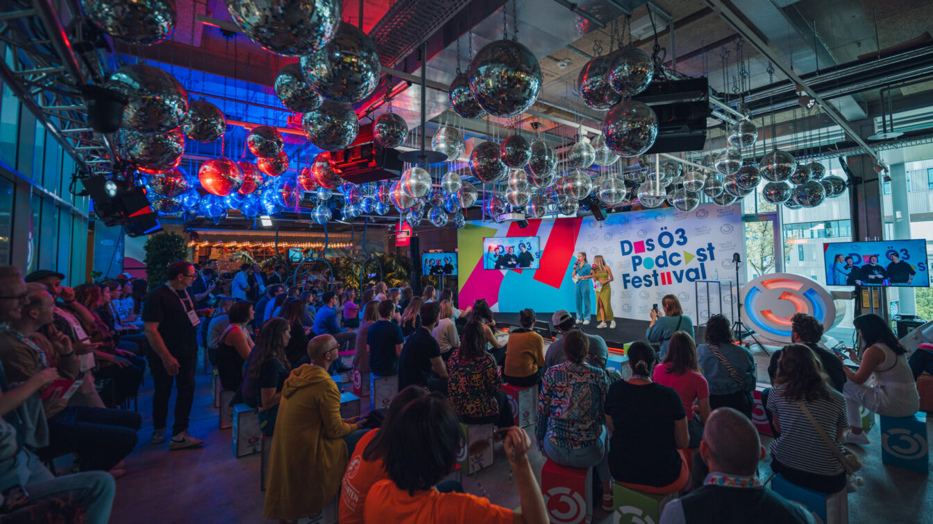 Das Ö3 Podcast Festival Bühne: Zwei Moderatorinnen vor einer weißen Wand mit buntem Veranstaltungslogo; rechts ein großes, beleuchtetes Ö3-Logo. Zuschauerraum: Gut gefüllt, Publikum sitzt auf bunten Hockern. Moderne Event-Location mit industrieller Decke, dicht behängt mit zahlreichen spiegelnden Discokugeln.Stimmungsvolles blaues und rotes Licht.