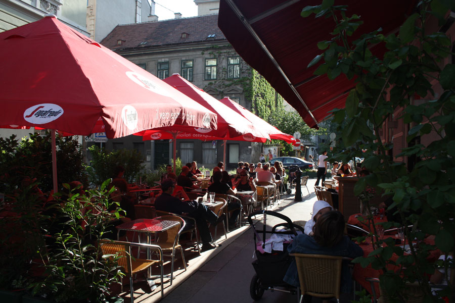 Tische und Sessel stehen unter Sonnenschirmen im Schatten im Gastgarten des Cafe Strozzi im 8. Bezirk in Wien