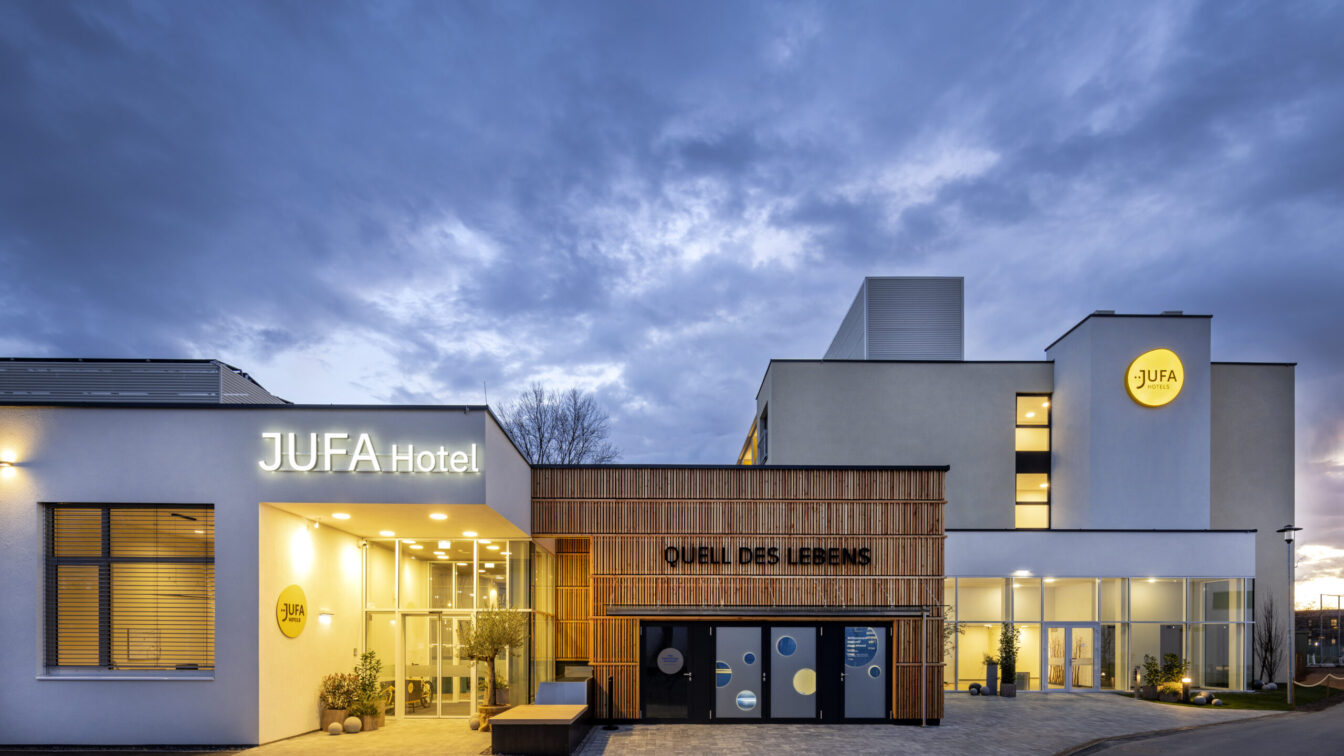 Sie sehen eine Außenansicht in der Abenddämmerung vom Haupt- und Nachteingang in das JUFA Hotel Bad Radkersburg sowie den interaktiven Erlebnisraum "Quell des Lebens" von außen.