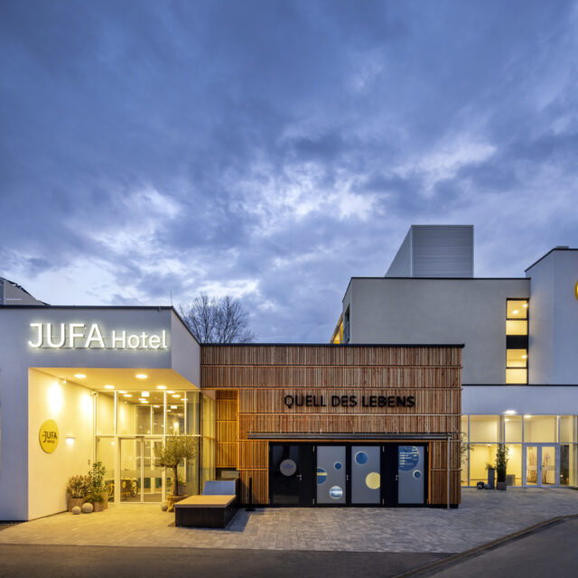 Sie sehen eine Außenansicht in der Abenddämmerung vom Haupt- und Nachteingang in das JUFA Hotel Bad Radkersburg sowie den interaktiven Erlebnisraum "Quell des Lebens" von außen.