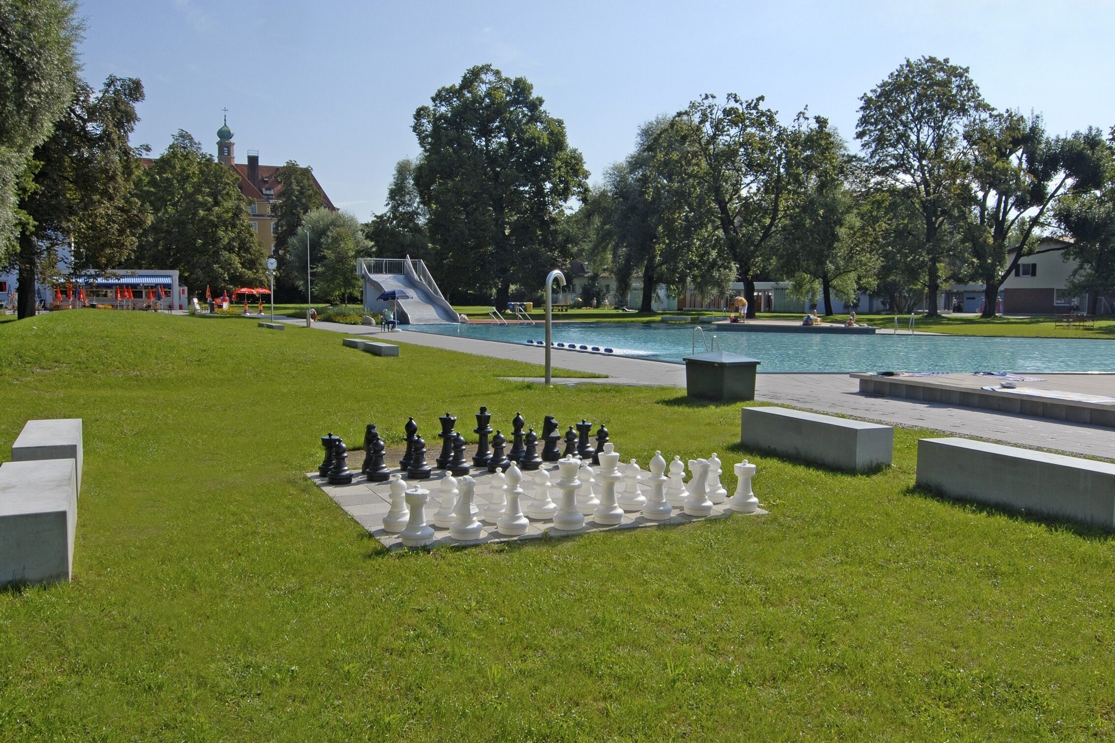 Ein Schachbrett mit großen Spielfiguren auf einer grünen Wiese mit Freibad in München im Hintergrund.